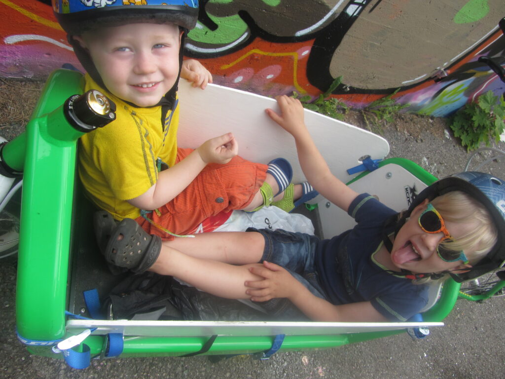 Två barn i en Bullitt lastcykel. De sitter å flakcykelns flak och ler mot kameran. De sitter inte fastspända utan sitter löst i lådan.